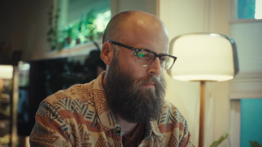 A concentrated bearded man with glasses works on his laptop in a cozy home office, with the screen reflecting in his lenses