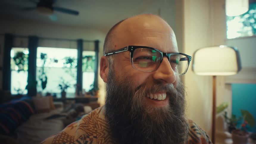 A bearded man with glasses smiles while working from home on his laptop in a cozy, plant-filled living room as the camera slowly pushes in