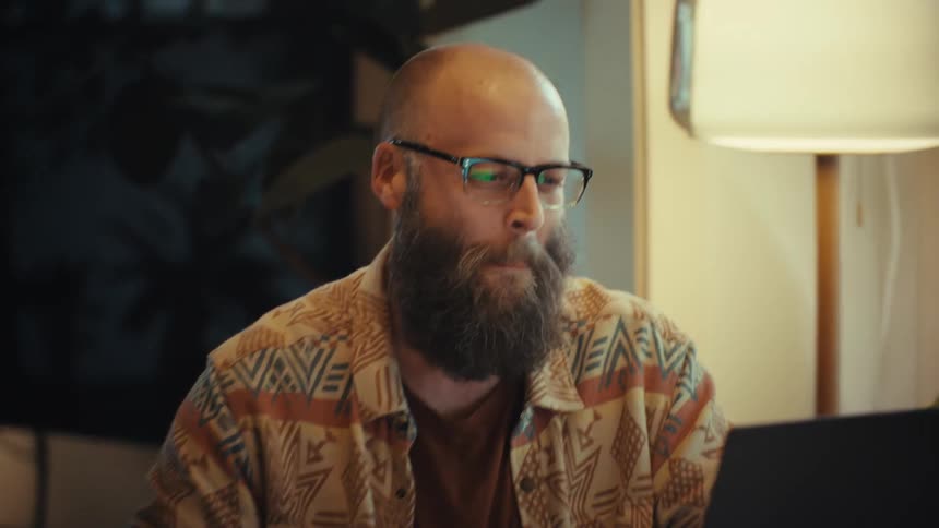 A happy bearded man wearing glasses enjoys a video call on his laptop while working from home in his cozy living room