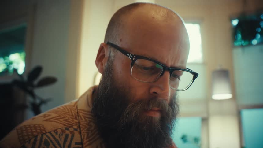 A focused, bearded man with glasses intently studies documents and takes notes at his desk in a home office