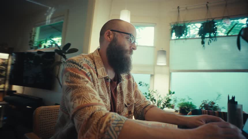 A bearded man wearing glasses diligently works on his laptop from the comfort of his cozy, plant-filled home office