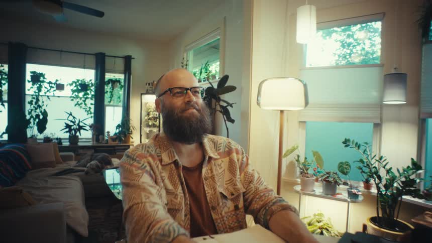 A bearded man working from his stylish, plant-filled home pauses from typing on his laptop to smile thoughtfully, as the camera slowly pushes in for a close-up