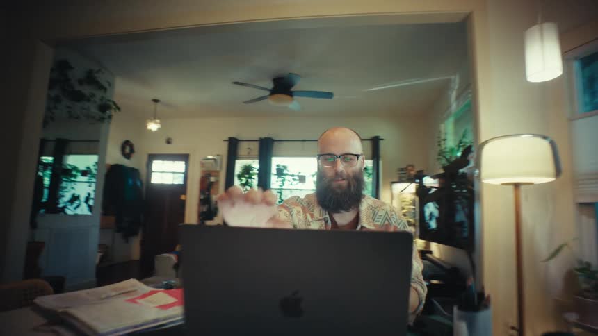 A stylish bearded man smiles with satisfaction after finishing his work on a laptop in his modern, plant-filled home as the camera pushes in