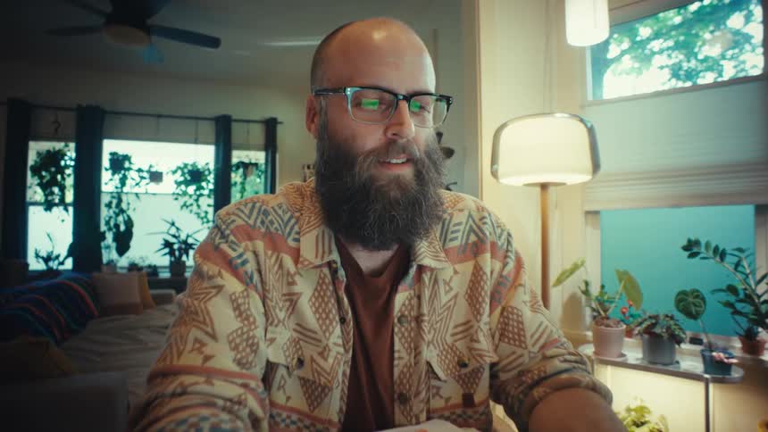 A stylish bearded man in glasses focuses on his laptop while working from his cozy, plant-filled home