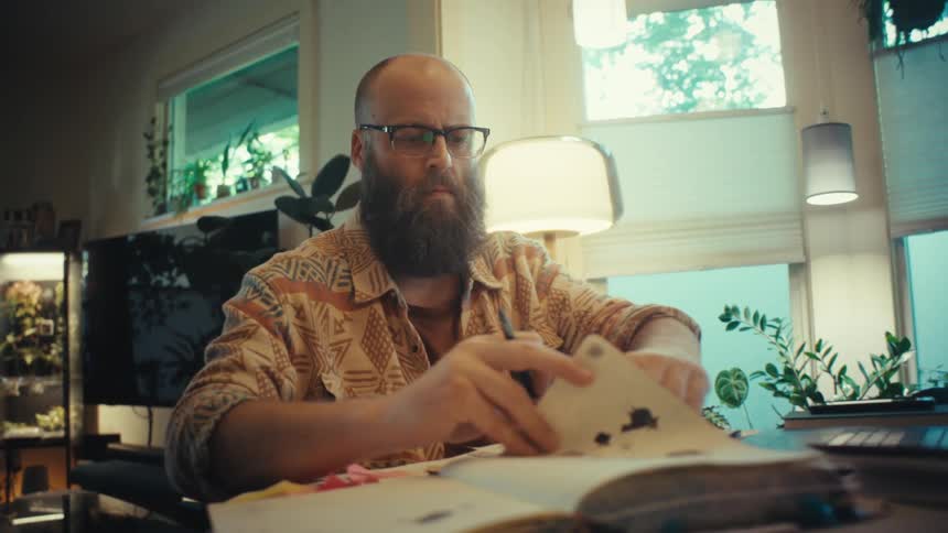 A focused, bearded man with glasses works from his home office, referencing his laptop, notes, and a sketchbook