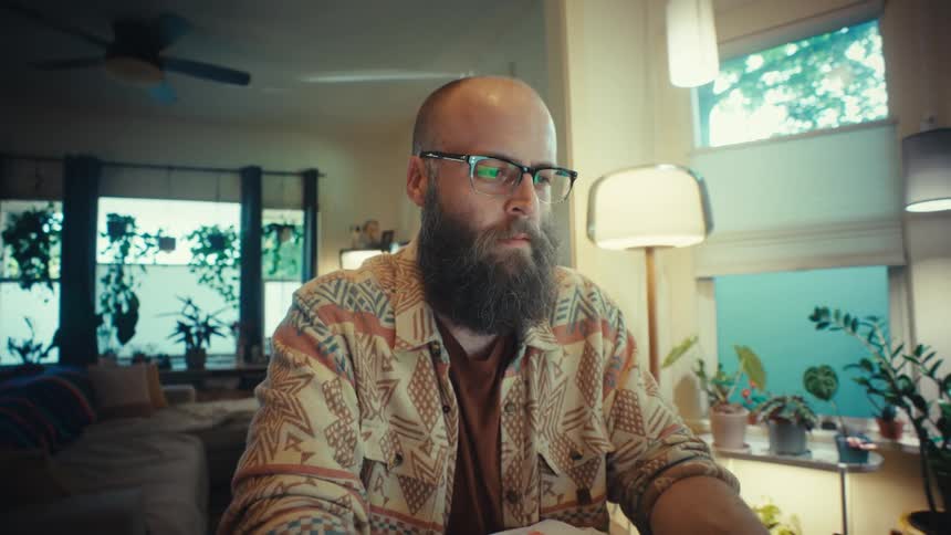 A bearded man with glasses works intently on his laptop from his home office in a slow dolly out shot