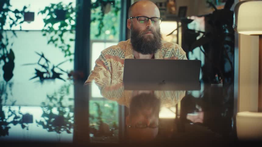 A bearded man in glasses works intently on his laptop at a reflective desk in a modern, plant-filled home office