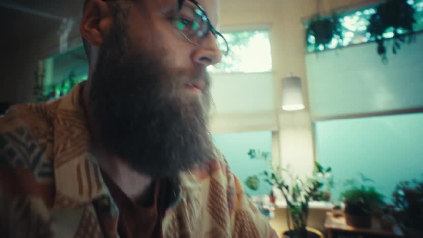 Close-up cinematic shot of a focused bearded man in glasses working from his sunlit home office, typing on a laptop at a cluttered desk
