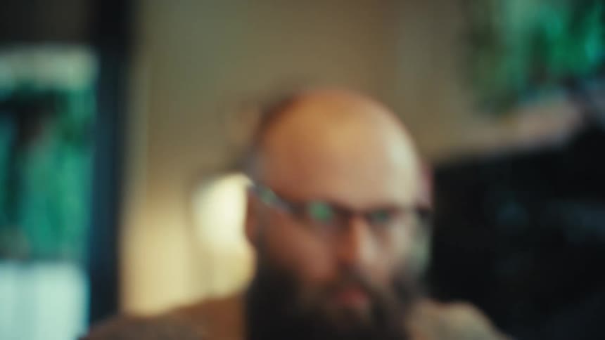 A bearded man in glasses wearing a patterned shirt walks to his desk and begins working on his laptop at home