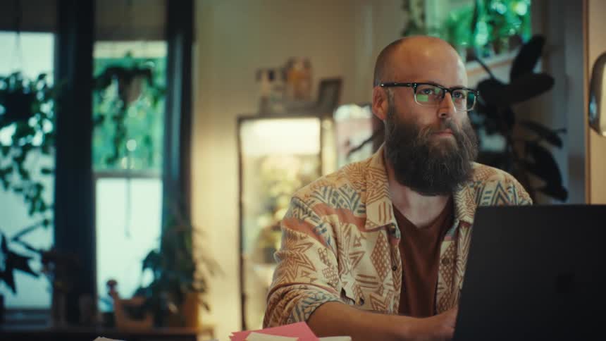 A man with a beard and glasses works on his laptop in a cozy home office filled with plants