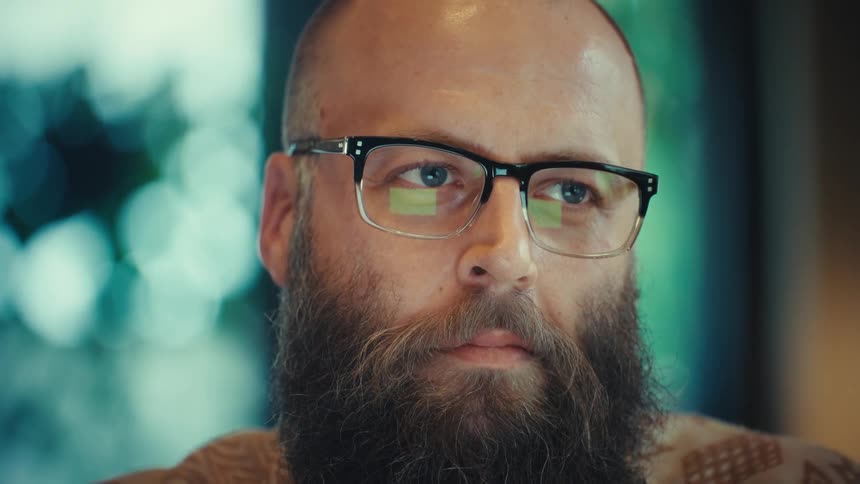 A focused bearded man wearing glasses works intently on his laptop, with the camera slowly zooming in on his thoughtful expression