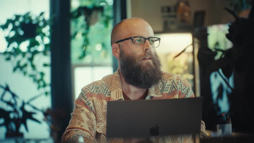 A pensive, bearded man wearing glasses works remotely on his laptop from a stylish, plant-filled home office