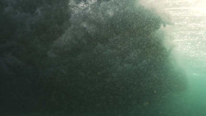 An immense ocean wave builds and crashes in dramatic slow motion, viewed from a turbulent underwater perspective with sunlight piercing the surface