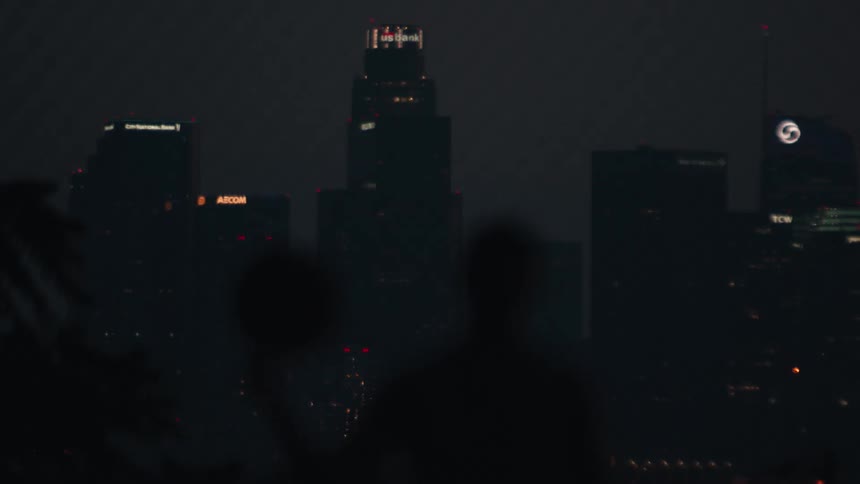 A hazy view of the downtown Los Angeles skyline at night, with the silhouettes of two people in the foreground