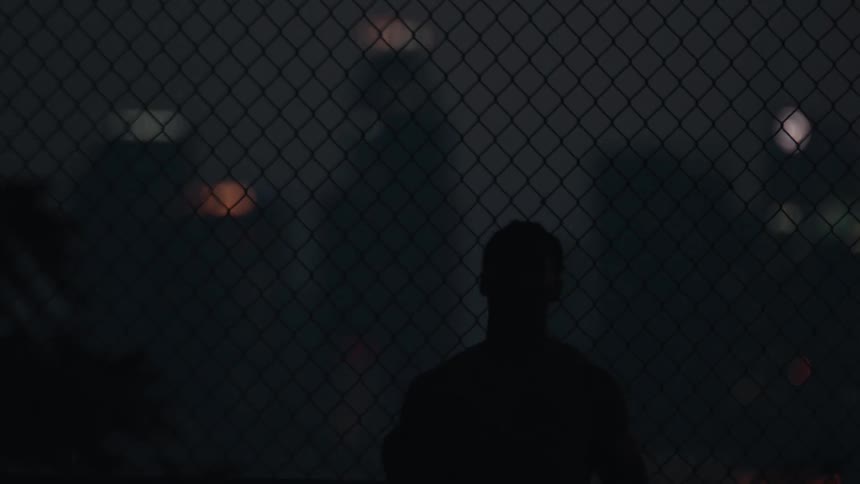 Silhouette of a man bouncing a basketball in front of a chain-link fence with blurry city lights in the background at night