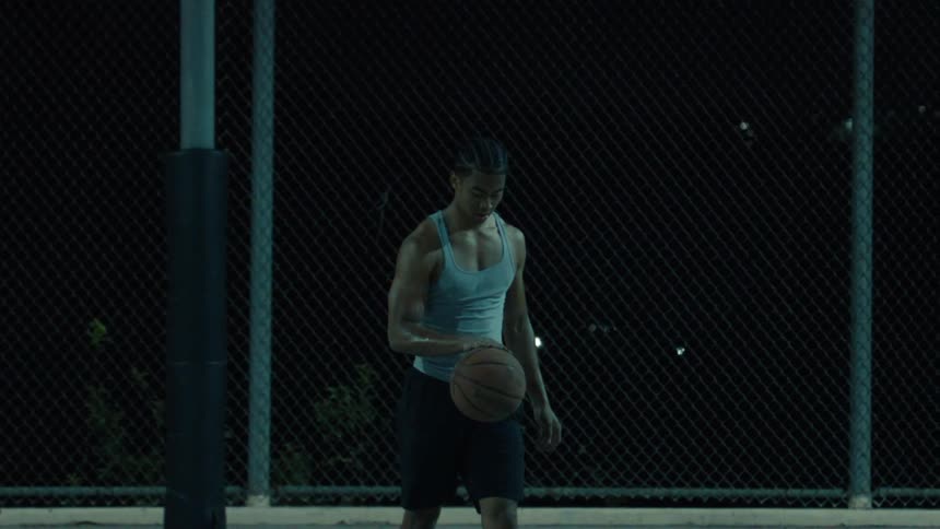 A muscular athlete practices basketball on an outdoor court at night, dribbling and spinning the ball in slow motion