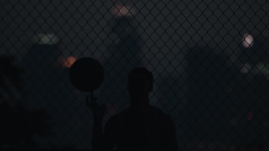 A slow-motion silhouette of a basketball player spinning the ball on their finger at night, framed by a chain-link fence with city lights in the distance