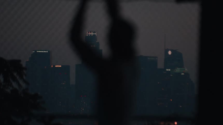 A contemplative silhouette of a person slowly moves in front of a window overlooking the Los Angeles skyline at dusk