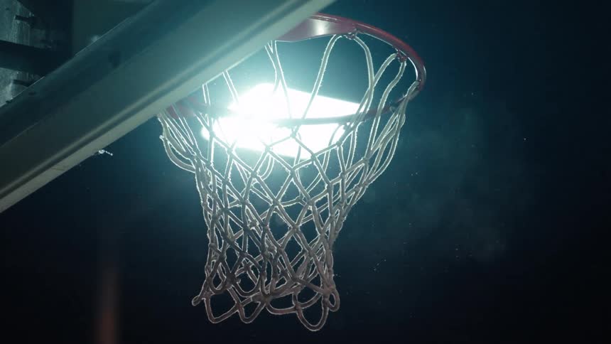 A basketball player dunks a ball in slow motion on a court at night, backlit by a bright stadium light