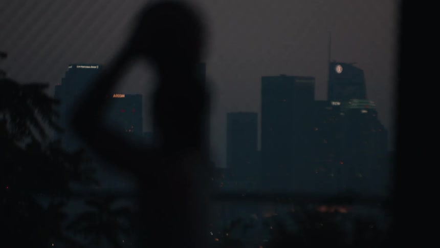 A silhouetted woman pulls her hair back while looking out over the hazy Los Angeles skyline at dusk