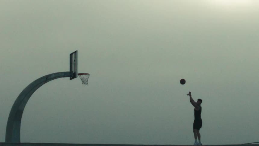 A silhouetted man shoots a basketball in dramatic super slow motion against a gray sky