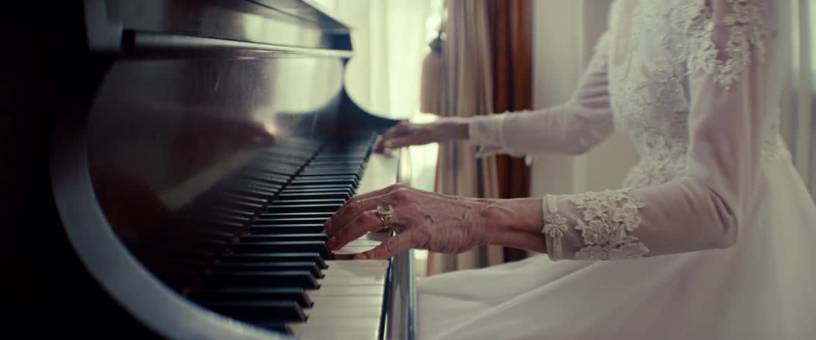 An elderly woman in an elegant white lace dress plays a classic wooden piano in a softly lit room