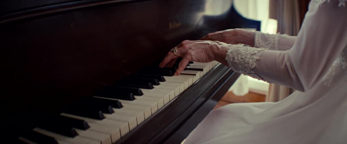 The graceful, aged hands of an elderly woman in a white lace dress play a gentle melody on a vintage piano