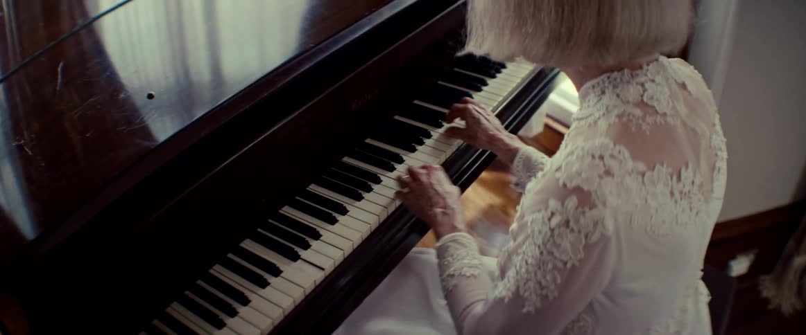 An elderly woman in a beautiful white lace dress gracefully plays a song on a wooden piano