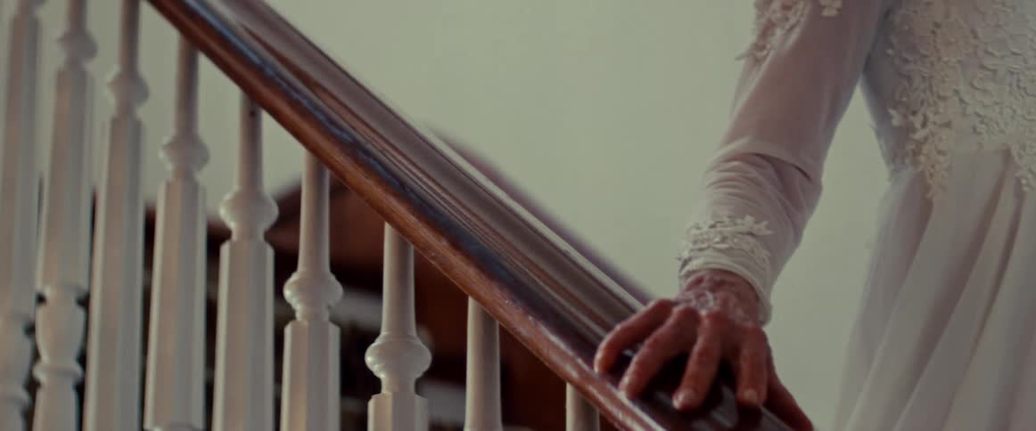 An elderly woman's hand slowly glides down a wooden banister as she descends the stairs in a white gown