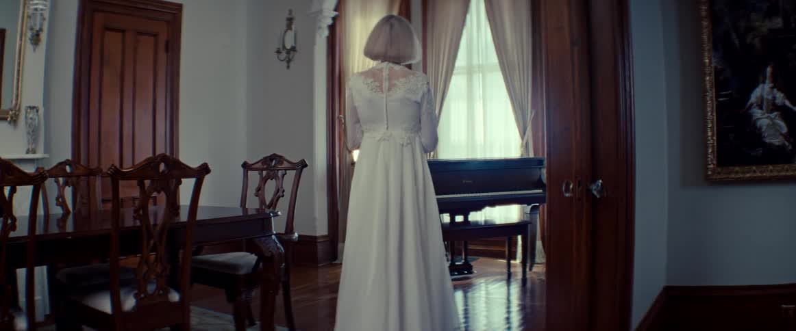 A bride in a vintage white wedding dress slowly walks through a dimly lit, elegant room towards a grand piano