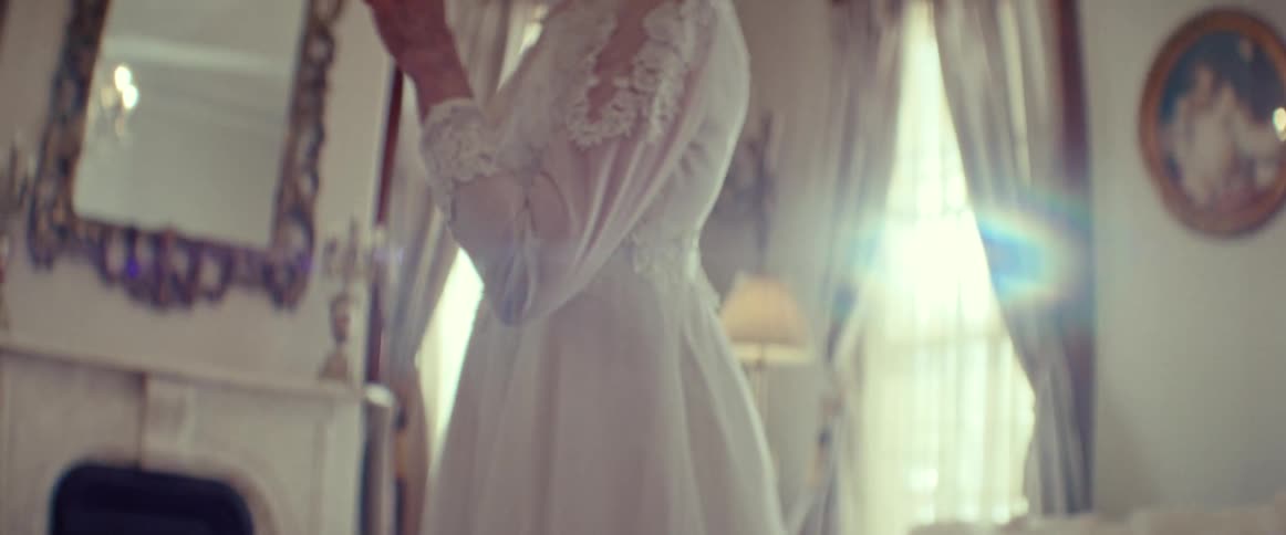 A nostalgic elderly woman smiles peacefully while trying on her vintage long-sleeved lace wedding dress in a sunlit room