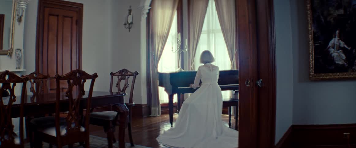 A woman in an elegant white lace gown plays a grand piano in a sunlit room of a stately home