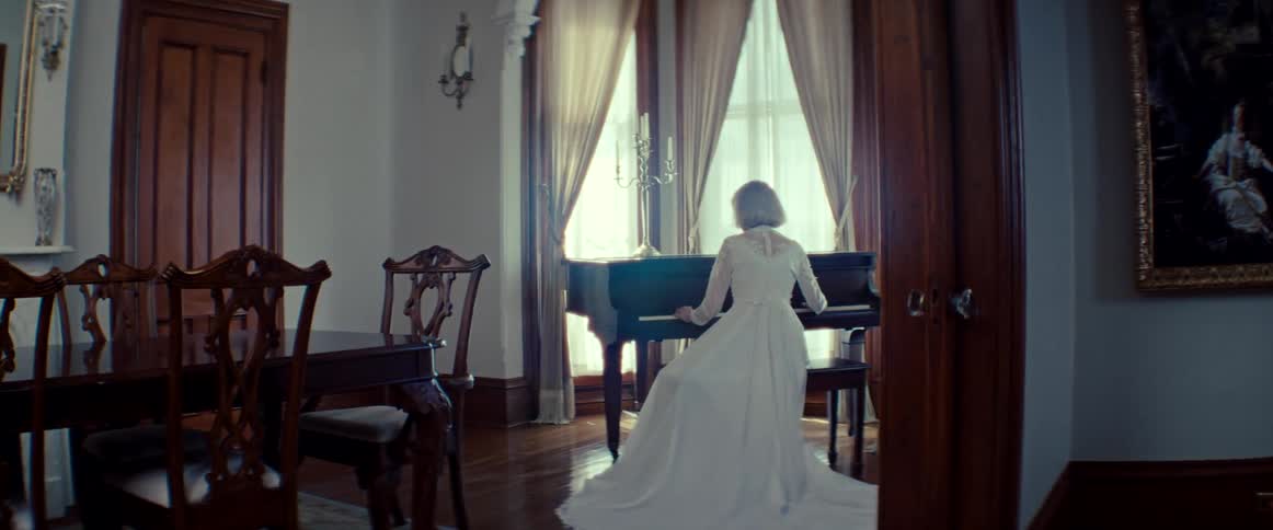 From behind, a woman in a long white dress plays a piano in a dark, antique room