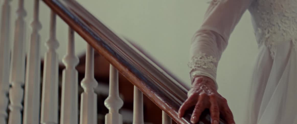 A close-up of an elderly woman's wrinkled hand slowly sliding down a wooden banister as she descends the stairs