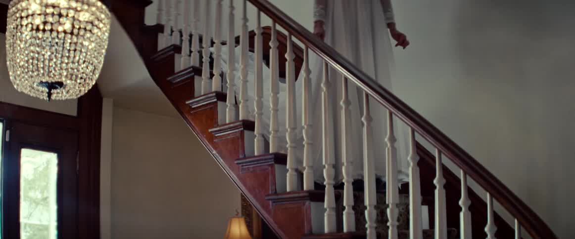 A ghostly figure of an older bride appears at the top of an ornate staircase in a dimly lit house