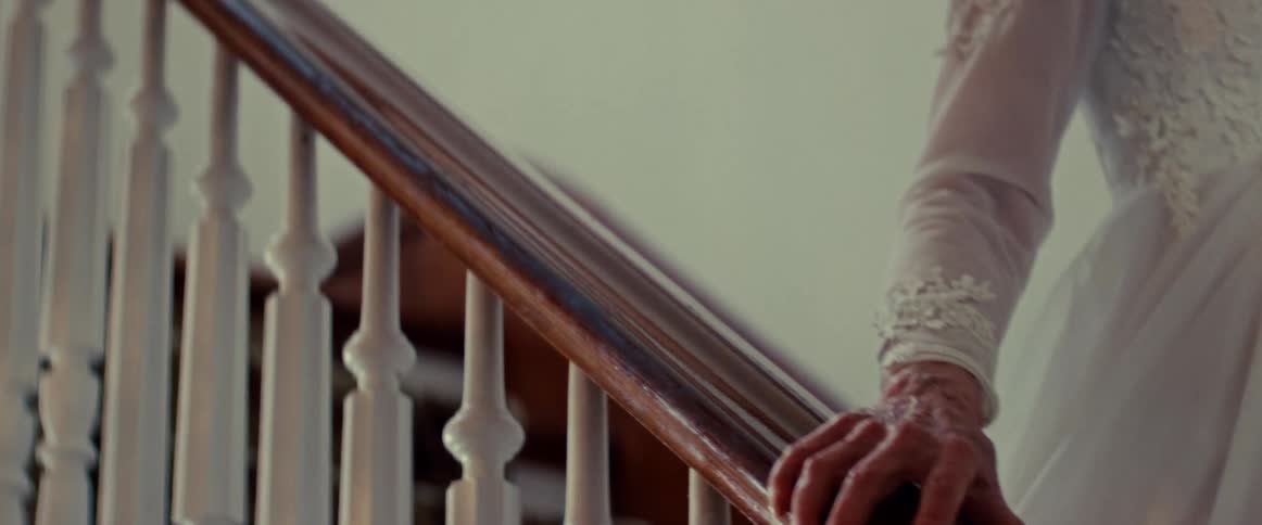 An elderly woman's wrinkled hand in a white lace dress slowly slides down a wooden staircase railing