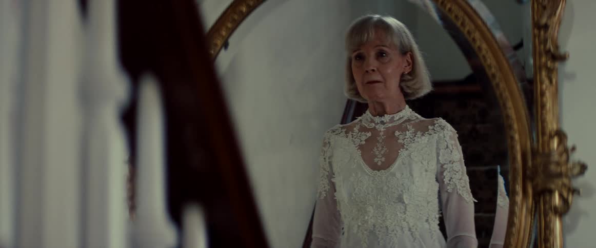 An elderly woman in a wedding dress smiles warmly at her reflection before it turns to a look of utter terror