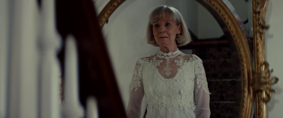 A senior bride in a beautiful white lace gown smiles happily at her reflection in an ornate vintage mirror