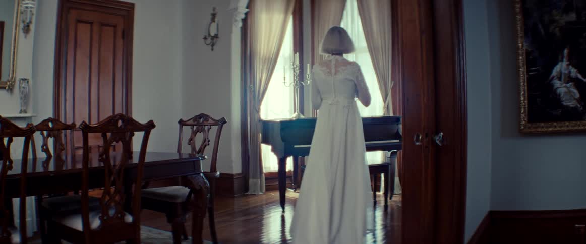 A woman in a vintage white gown gracefully walks towards a grand piano in a sunlit, classic room