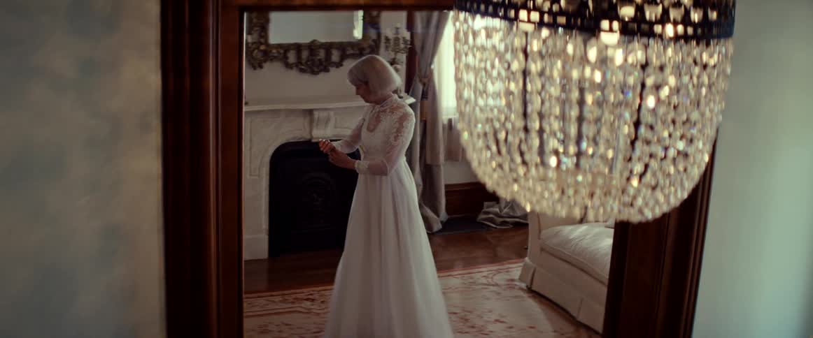 A senior woman in a beautiful white wedding dress gently twirls and admires the gown in a grand, vintage-style home