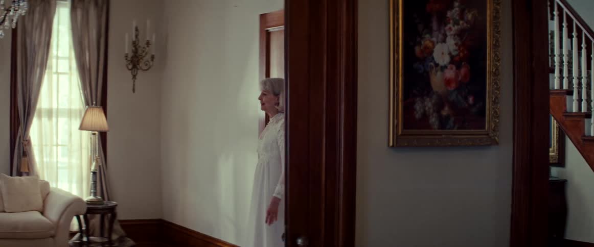 A smiling senior woman in a vintage white wedding dress walks from a classic staircase into the formal living room of her elegant home