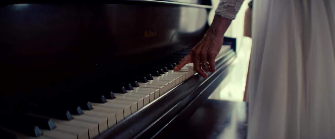 An elderly person's hand gently glides down the keys of a classic wooden piano in a softly lit room