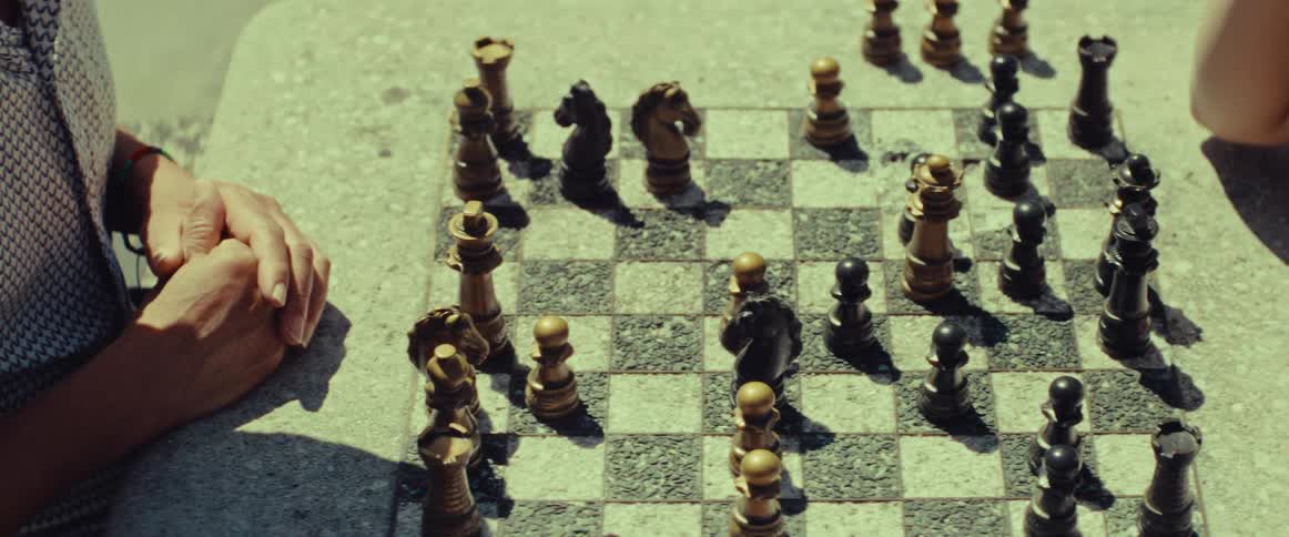 A high-angle view of a person's hands resting by a weathered stone chessboard during a sunny outdoor game
