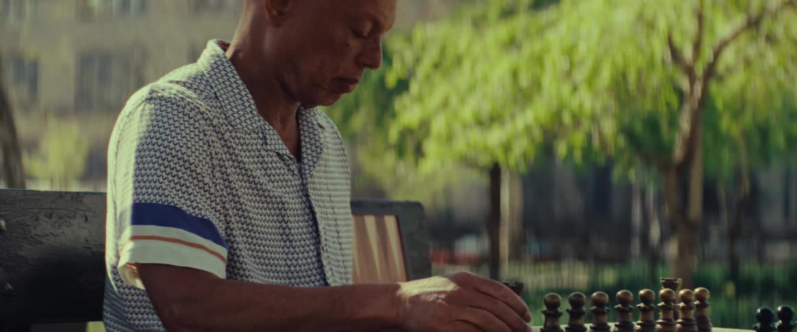 An elderly man thoughtfully plays a game of chess by himself at a stone table in a sunny park