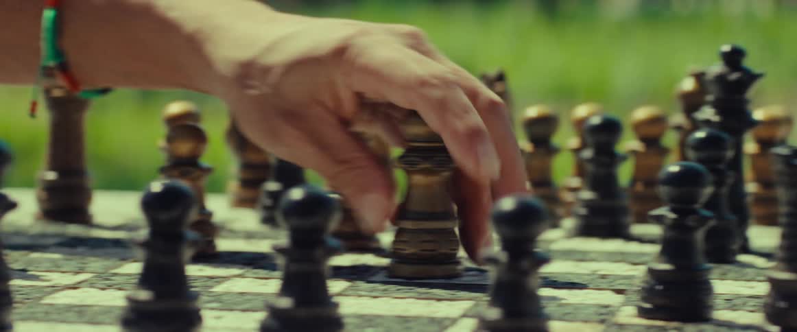 A person's hand makes a thoughtful move on a weathered chessboard in a sunlit outdoor setting, shown in slow motion
