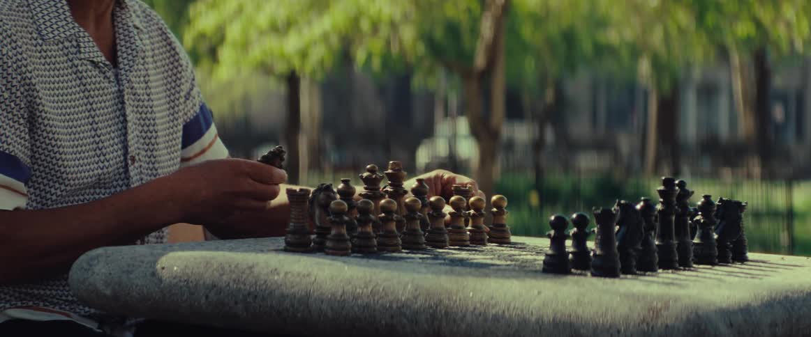 A senior man's hands set up wooden chess pieces on a stone table in a sunny park, preparing for a game