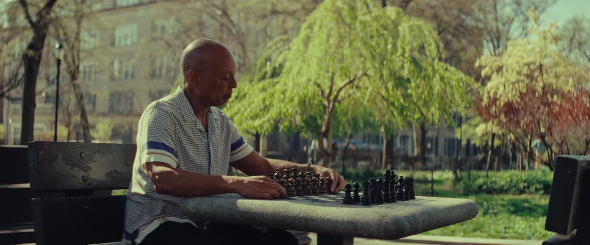 An older man thoughtfully sets up a chess game on a stone table in a sunlit urban park