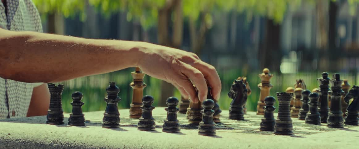 A close-up reveals a man's hand making a strategic move during a chess game on a stone table in a sunny park
