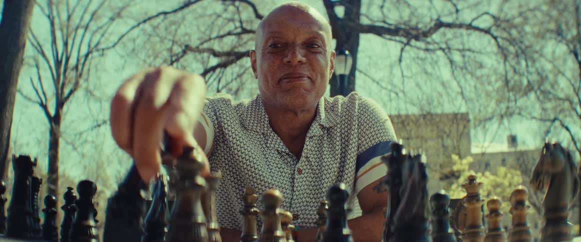 A confident older man smiles as he plays a strategic game of chess in a sunny park, seen from his opponent's perspective