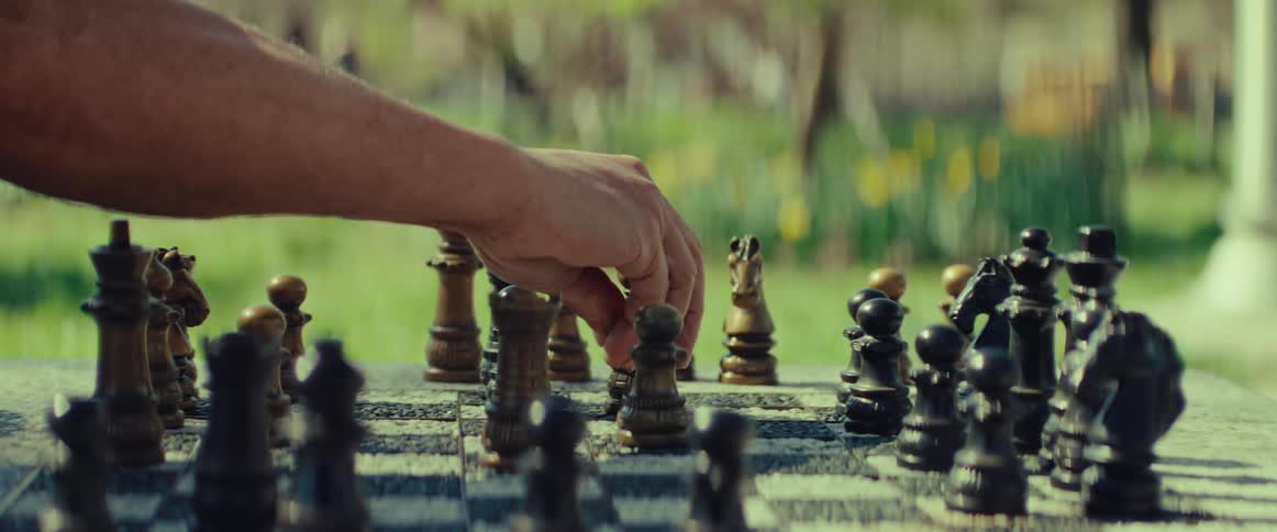 A close-up shot captures a strategic chess move being made on a stone table in a sunlit park